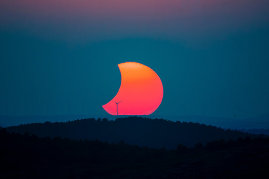 Partially eclipsed sun setting behind wind turbines and forested hills at dusk