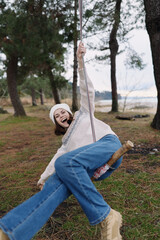 Obraz premium Joyful woman swinging outdoors in a forest park, laughing and leaning back on a rope seat, wearing a beanie, sweater and jeans. Vivid trees and grass create a carefree, candid moment of play.
