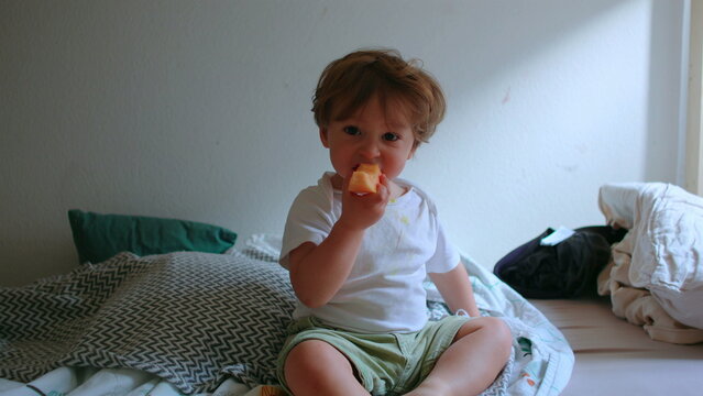 Toddler sitting on bed eating melon slice during relaxed snack time, looking toward camera with curious expression, healthy childhood nutrition and everyday family life at home