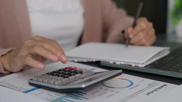 Business accounting concept showing woman hands using calculator and writing on notebook with budget report chart on desk. Ideal for financial planning, tax analysis and corporate audit media.