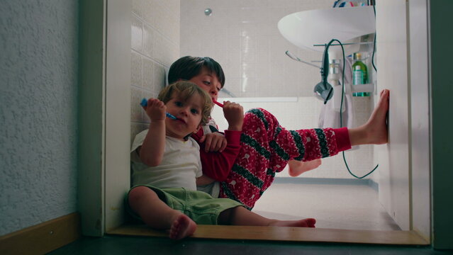 Young siblings sharing bedtime hygiene routine in bathroom while brushing teeth together creating authentic family moment centered on health responsibility and childhood learning