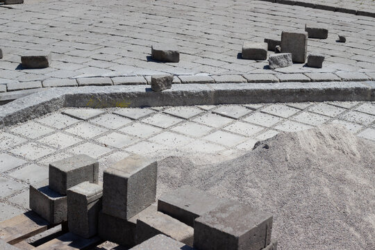 Construction site featuring gray paving stones, scattered stone blocks, and a pile of sand on a sunny day with a patterned stone surface in the background