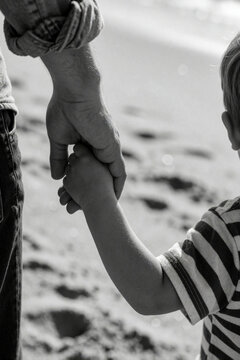 Father holds son hand on sandy beach shore. Man and boy walk together. Childhood memory, parental bond, family connection, love, safety, guidance.