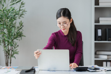 Focused Professional at Her Desk: A young Asian woman with glasses, dressed in a purple top, sits...