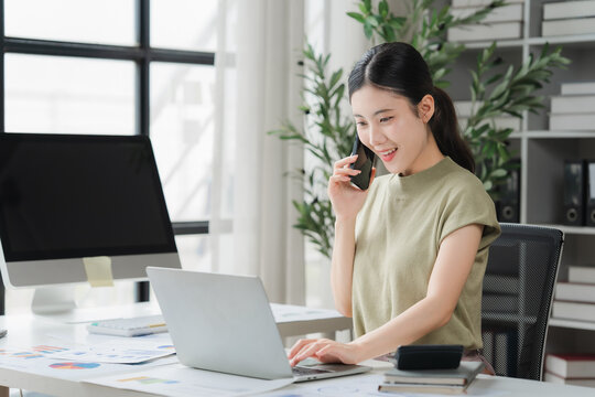 Focused Professional: A young Asian woman with a cheerful expression, engaged in a phone call while working on her laptop at a modern office desk.