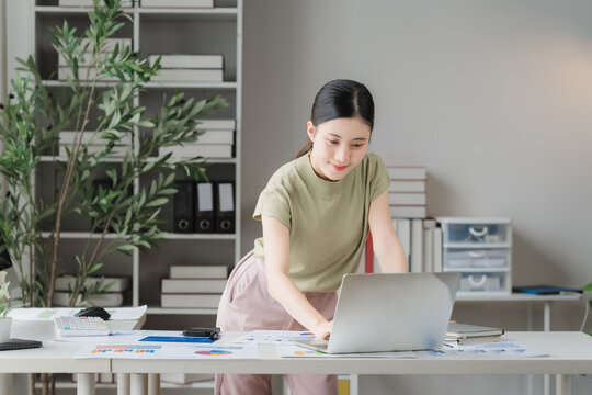 Young woman working at a desk in a home office, surrounded by plants and organized shelves. She is focused on her laptop, a picture of productive multitasking.