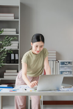 Focused Professionalism: A young woman diligently works on her laptop at a modern desk, surrounded by office supplies and stacks of books, embodying concentration and dedication in her task.