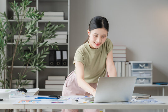 Focused Professional: A young woman works diligently on her laptop in a bright, organized office space, surrounded by plants and shelves filled with books and files.