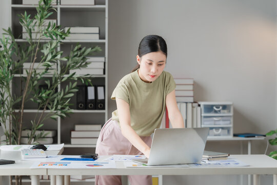 Focused Professional at Work: A young woman intently works on her laptop at a well-organized desk, surrounded by office supplies and a large bookshelf filled with books and binders.