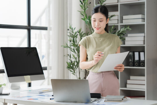 Young Professional Woman in Modern Office: A focused and professional young woman reviews documents in a bright, contemporary office space, surrounded by technology and organized shelving.