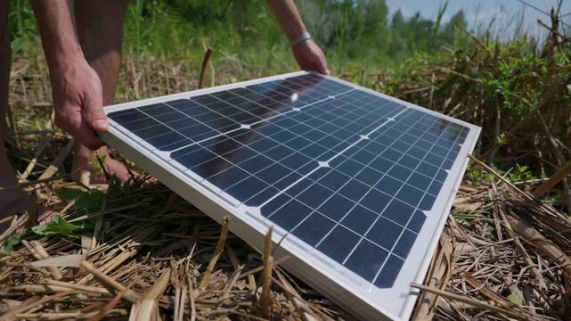 A person carefully places a portable photovoltaic solar panel on dry grass in nature. Concept of renewable green energy, eco-friendly lifestyle, off-grid living, and self-reliance.