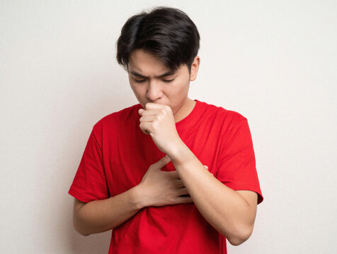 Young Asian man in red shirt coughs into hand. Sick guy feels chest pain discomfort suffering from flu symptoms. Male person respiratory illness, cold virus infection. Isolated studio shot on white
