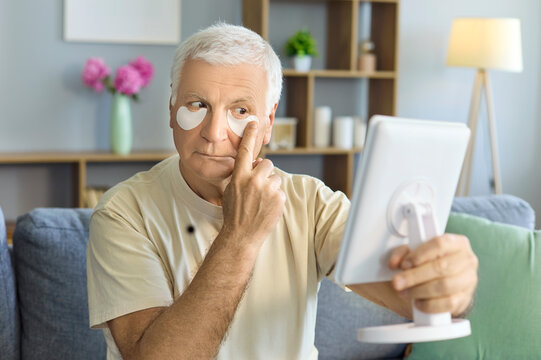 Senior man with patches, using mirror, home skincare. Under eye pads ease puffiness and circles as he perfects a face grooming routine, promoting aging and hygiene. Concept of male eye care.
