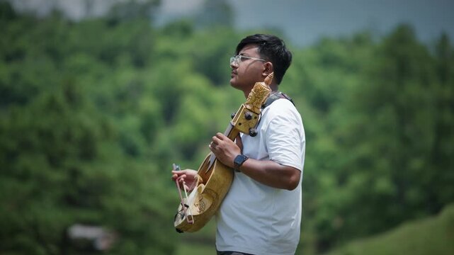 Young man in white shirt plays a hand carved sarangi with carved wooden head visible against a softly blurred green forest background