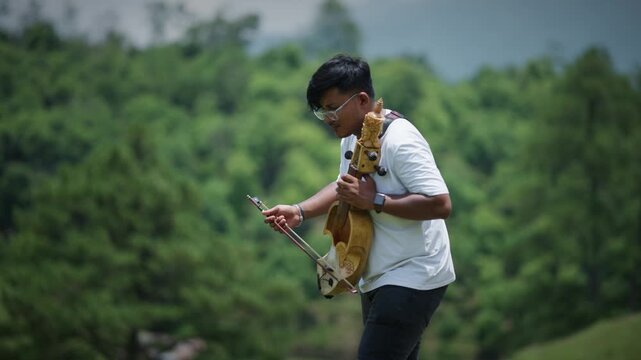 Side profile of a young man in white shirt playing a traditional sarangi outdoors with soft green forest bokeh blurred in the background