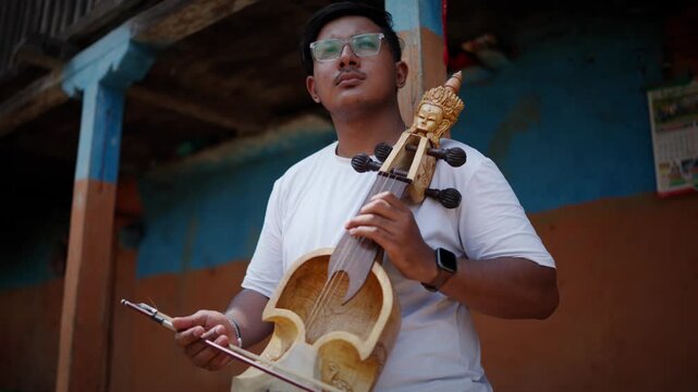 Low angle portrait of a young man wearing a cap and glasses holding a carved sarangi in front of a traditional wooden balcony house