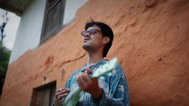 Low angle portrait of a young man wearing sunglasses playing a small ukulele against a weathered orange mud wall of a traditional village house