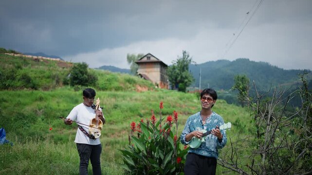 Two musicians play ukulele and sarangi among red flowering plants on a hillside with a traditional wooden house visible under a dramatic cloudy sky