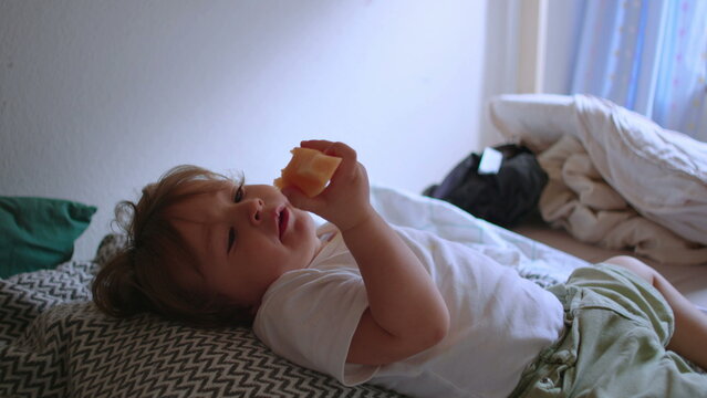 Toddler enjoying melon snack while sitting comfortably on bed, peaceful home scene focused on healthy eating, childhood independence and everyday family routine