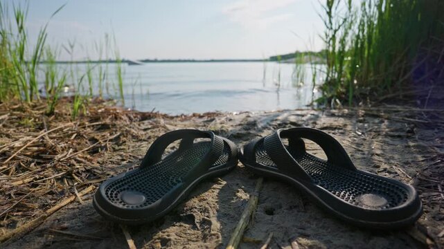 Black sandals rest on the sandy shore surrounded by wild green reeds near calm lake water. Concept of summer vacation, outdoor relaxation, beach lifestyle, and casual holiday freedom.