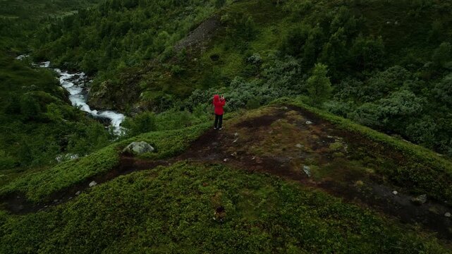 Individual in red jacket standing on a hilltop overlooking a flowing river surrounded by lush greenery and rocky terrain in a mountainous landscape