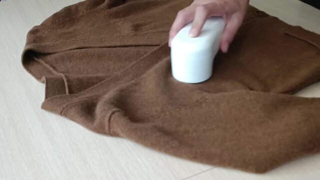 A close up of a hand using a white fabric shaver to remove lint and pilling from a brown sweater on a light wooden surface