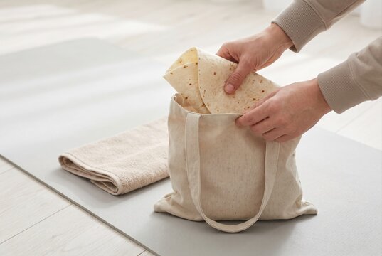 Hands packing rolled flatbreads into a reusable canvas tote bag on a fitness mat