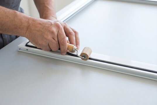 Worker hands using a wooden roller tool to press black rubber spline into a white window screen frame