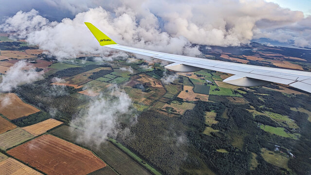 Scenic aerial view from airplane window during daytime flight, with airBaltic aircraft wing, patchwork fields and forest landscape: Riga, Latvia - 17 August, 2025