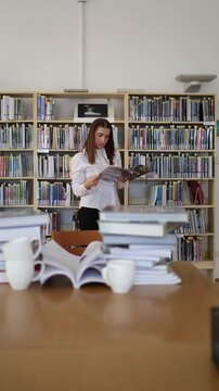 Vertical screen young female student reads a book while searching for study material among library shelves surrounded by stacks of books during a focused exam preparation