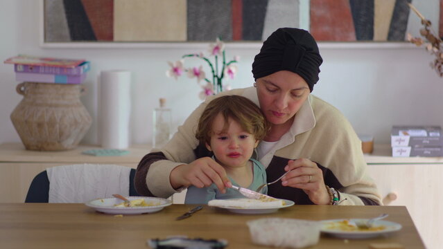 Mother eating meal while toddler rests on her lap at dining table in family home during cancer recovery, highlighting caregiving, resilience, emotional support and family connection