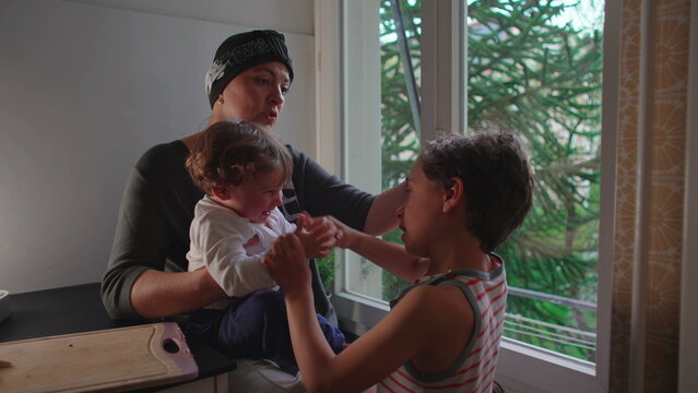 Mother caressing older son while he playfully claps toddler&rsquo;s hands together, authentic family interaction expressing affection, sibling bonding, parental love and emotional connection at home