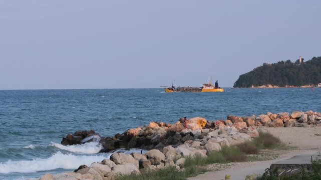 Dredge ship sailing near a rocky coastline at sunset