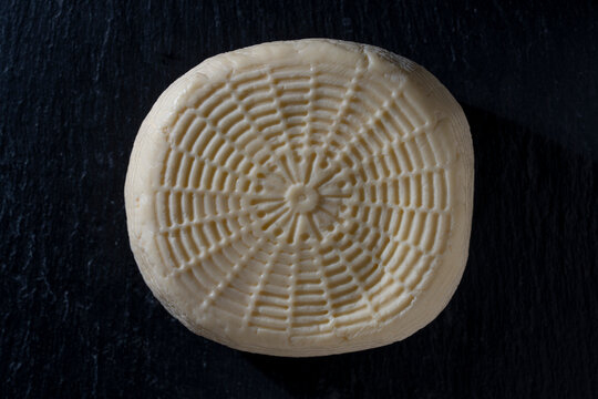 Whole round head of homemade cheese on dark background, closeup. Top view of fresh whole round of artisanal soft cheese or sulguni showing basket mold wicker texture placed on black slate surface