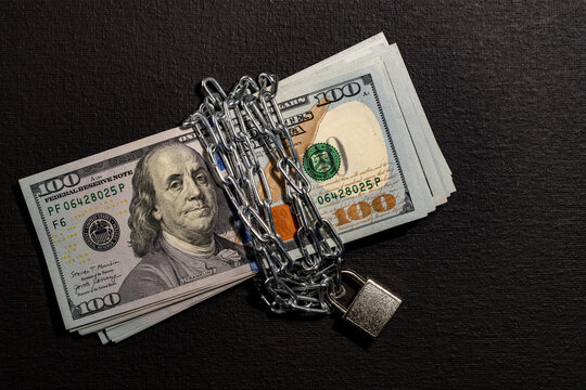 US dollar bills wrapped in chain with padlock, closeup. Stack of one hundred dollar cash notes securely bound with steel chain and locked padlock on dark black textured background
