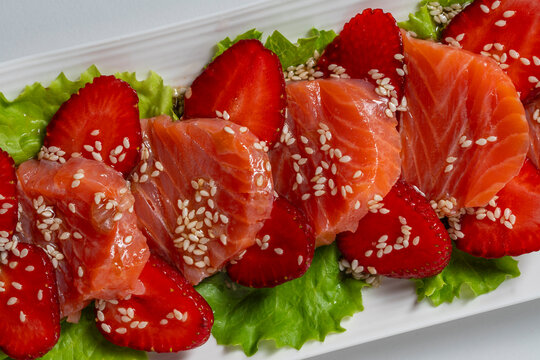 Salmon sashimi with fresh strawberries. Closeup of gourmet salmon carpaccio paired with red strawberry slices on green lettuce sprinkled with sesame seeds filling the frame in food styling concept