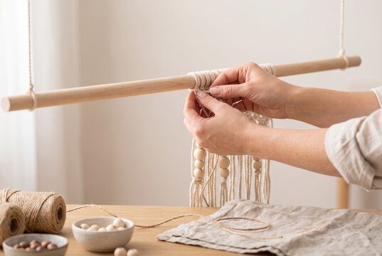 Hands tying thick cotton macrame cords onto a wooden dowel rod to create a handmade wall hanging