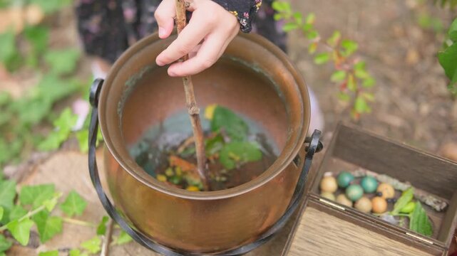 child hand stirring copper cauldron with stick, natural ingredients, water with leaves and berries inside, Halloween or fantasy witch crafting setup, Esoteric practice herbal medicine and magic potion