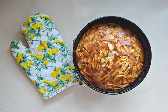 Homemade apple sponge cake, traditional apple sharlotka, with baked apple pieces and golden crust in round pan beside oven mitt on light kitchen surface, top view