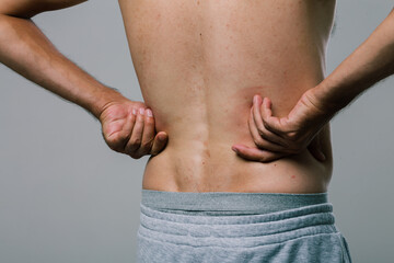 Naklejka premium A man with a summer tan massages his sore lower back with his fingers. A guy in gray shorts, turning away from the camera, scratches his pimply back. men's health: prevention of spinal diseases
