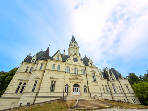 Castle towers under the sky. A large castle is seen in the background with trees around it. The sky is bright and cloudy, creating a historic view.
