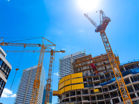 Construction occurs on-site. Heavy machinery and cranes work on a tall building under a bright sky with no clouds.