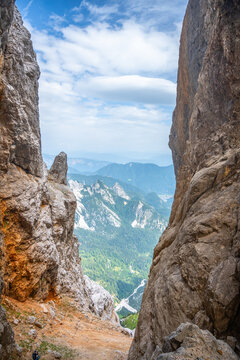 The scene shows a gap between two large rock formations in the Julian Alps. The view reveals green valleys and distant mountains under a cloudy sky. A person can be seen in the distance.