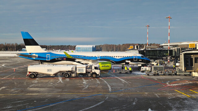 AirBaltic passenger aircraft during ground handling at Riga International Airport with baggage carts, fuel truck, workers and jet bridge on winter apron: Riga, Latvia - January 13, 2025