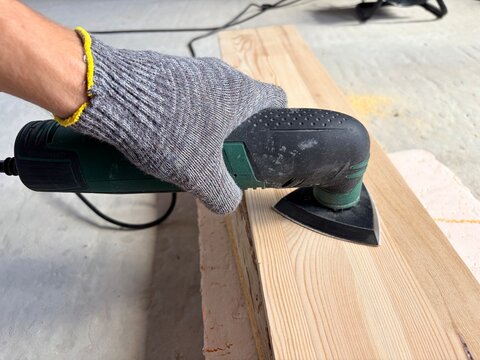 a worker's gloved hand using an electric detail sander on a wooden plank