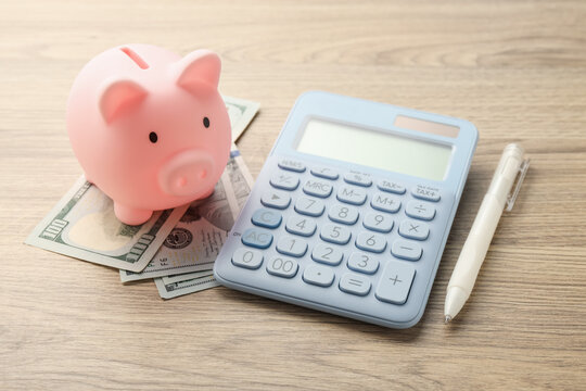 Piggy bank, calculator, dollar banknotes and pen on wooden table, closeup