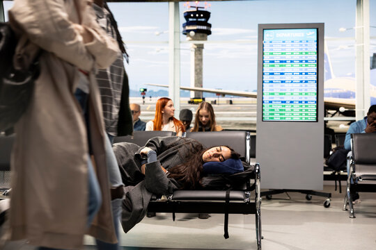 Exhausted woman sleeps across metal chairs in a bright airport terminal waiting area during a long flight delay and international journey, resting until departure resumes for her next flight.