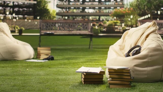 Wireless headphones rest on a fabric beanbag outdoors, framed as technology detail with stacked books, greenery and modern buildings on campus blurred behind, reflecting student life.