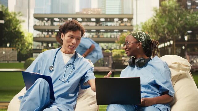 Medical students in blue scrubs compare notes during a relaxed outdoor group study, sitting on beanbags and discussing healthcare, with a laptop and open book serving as study materials.