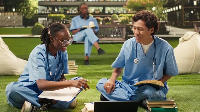 Diverse medical students make a fist-bump gesture beside an open laptop on grass, with friends celebrating progress and teamwork after collaborative learning outdoors, during a study session.